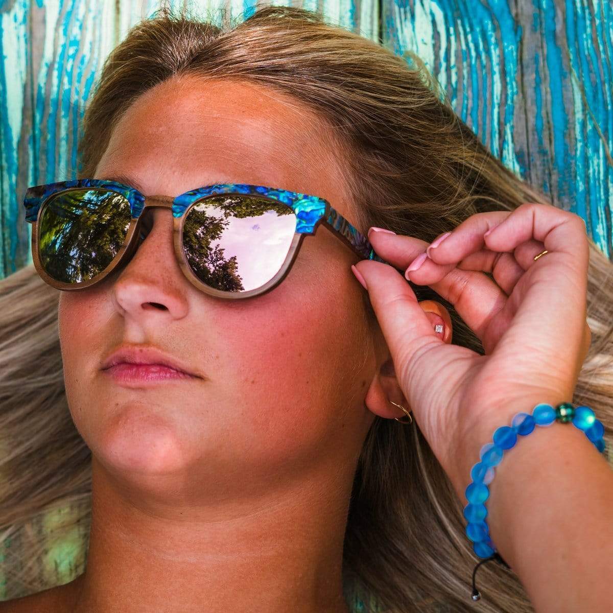 Model Tanning Outside Is Wearing Beachcomber Abalone Wooden Sunglasses With Rose Lenses